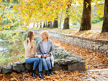 Eine jüngere Frau sitzt mit einer älteren Frau auf einer niedrigen Steinmauer. Sie trinken Kaffee aus einem Becher.