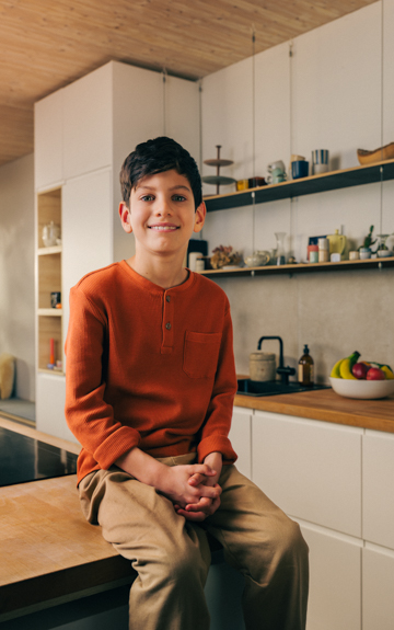Ein kleiner Junge mit Locken lacht in die Kamera und hält ein gelbes Skateboard hinter seinem Rücken fest.