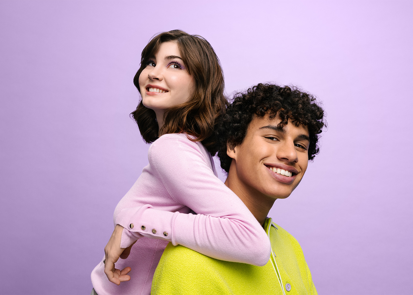 Eine junge Frau lehnt sich mit Rücken und Ellenbogen auf die Schultern eines jungen Mannes. Beide lächeln.