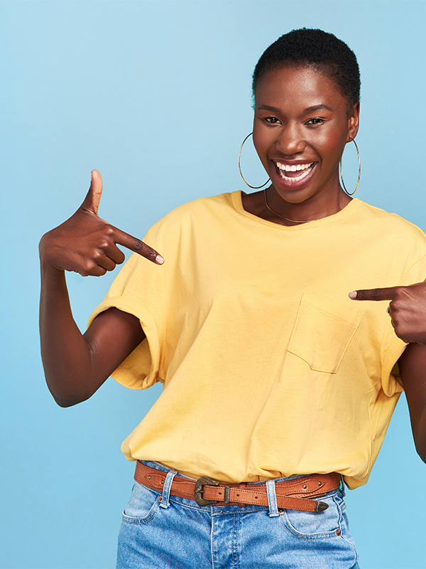 Eine lachende Frau steht vor hellblauem Hintergrund und zeigt mit beiden Zeigefingern in die Kamera.